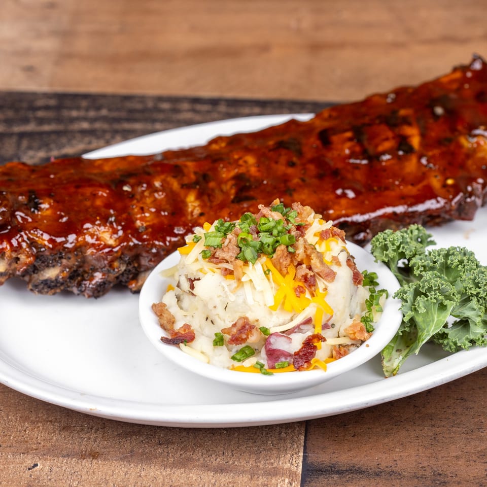 A rack of BBQ ribs with a side of loaded mashed potatoes and kale on a white plate.