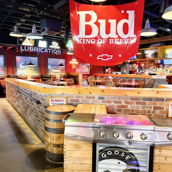 A rustic restaurant interior with brick walls, wooden tables, and a large Budweiser sign hanging overhead.
