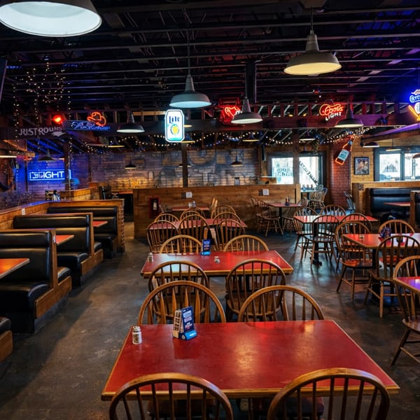 Spacious, dimly lit restaurant interior with red-topped tables, wooden chairs, booths, and neon signs.