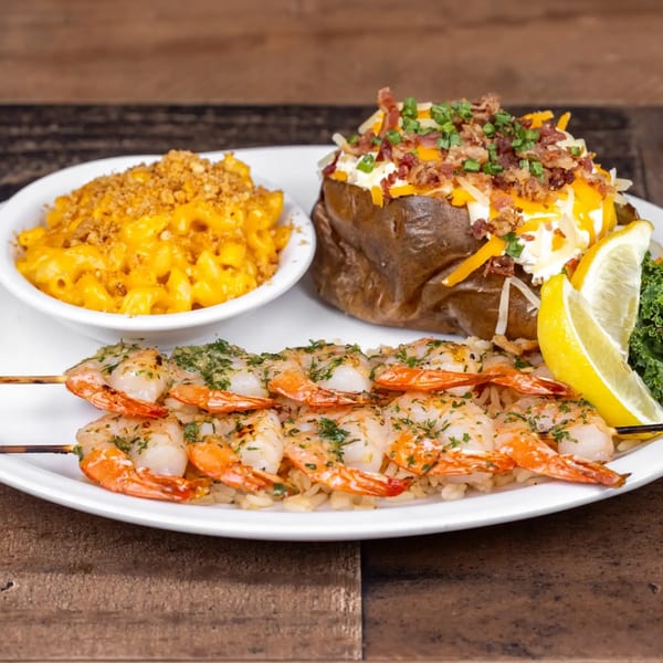 Grilled shrimp skewers on rice, with a side of mac and cheese and a loaded baked potato.