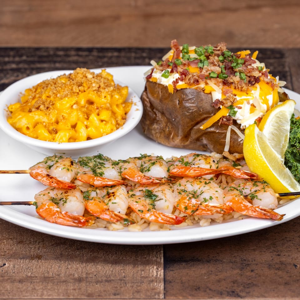 Grilled shrimp skewers on rice, with a loaded baked potato and mac and cheese.