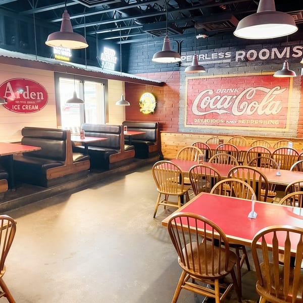 A retro-themed restaurant interior with red tables, wooden chairs, booths, and vintage Coca-Cola and Arden Ice Cream signs.