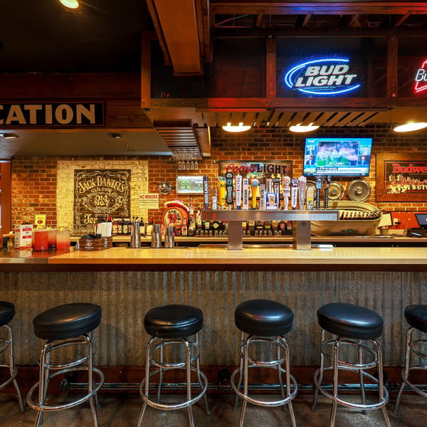 A rustic bar with brick walls, neon signs, beer taps, and black bar stools.