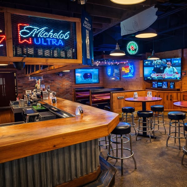 A rustic bar with a wooden counter, neon signs, TVs, and high-top tables with stools.